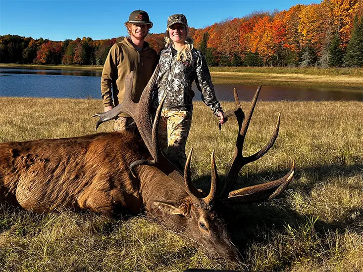 A man and a woman standing next to an elk in front of a beautiful lake in autumn